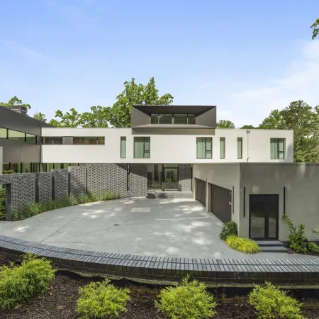 Modern luxury house with white and gray exterior, large driveway, and surrounding greenery under blue sky.