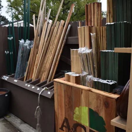 Stacks of wooden stakes and green metal garden poles stored outdoors with a wooden crate labeled AG for gardening supplies.