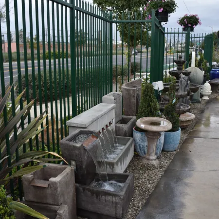 Outdoor garden area with stone fountains, planters, and green metal fence under cloudy sky.