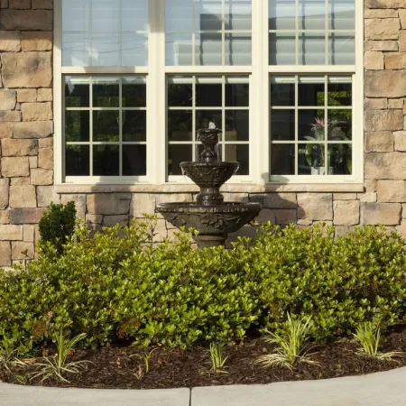 Stone house wall with large windows, central black tiered fountain, green shrubbery, and flower pots on stone pillars.