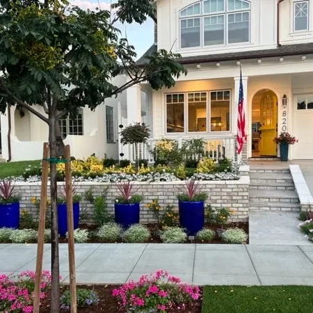 Suburban house with white brick wall, blue planters, American flag, and colorful flower garden in front yard.