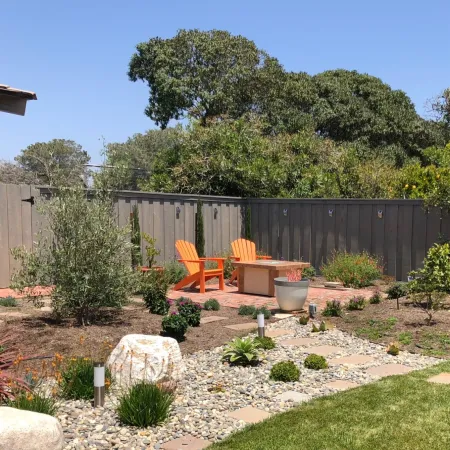 Backyard garden with orange chairs, fire pit, stone pathway, and various plants under a bright sky.