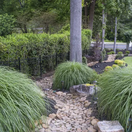 Stone pathway with ornamental grasses, large rocks, black metal fence, and manicured garden landscaping.