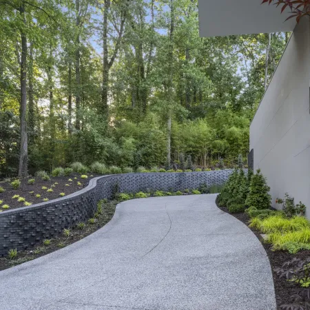Curved concrete driveway bordered by landscaped garden and retaining wall beside modern house in wooded area.