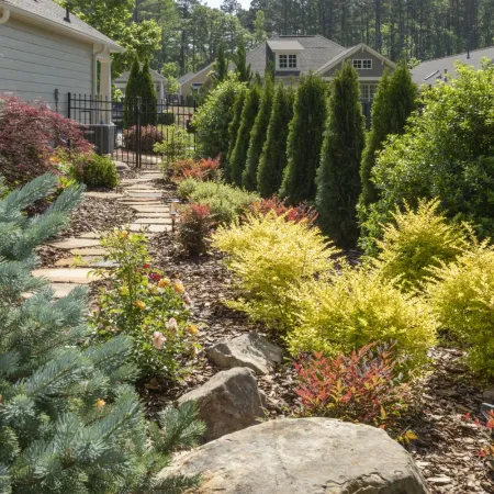 Garden pathway lined with colorful shrubs, evergreen trees, and decorative rocks beside a residential house