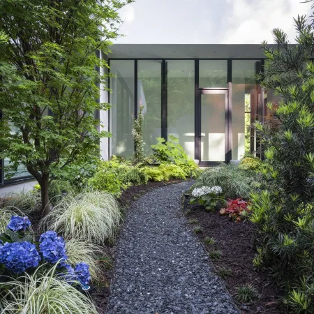 Gravel path leading to a modern glass door surrounded by lush green plants and colorful flowers.