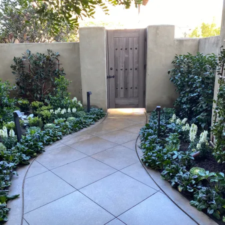 Curved stone pathway through lush garden beds leading to a closed wooden gate in a courtyard.