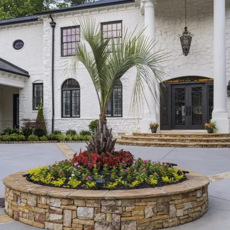 Stone-bordered circular garden bed with palm tree and flowers in front of white house with columns and black doors.