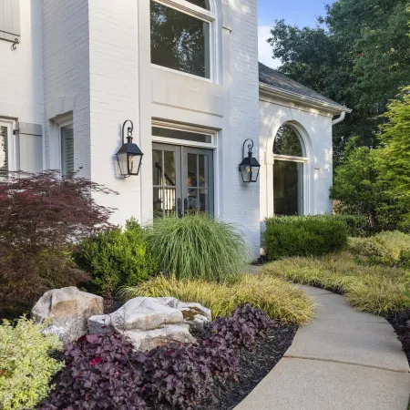 Curved garden path leads to white house entrance surrounded by diverse green shrubs, plants, and decorative rocks.