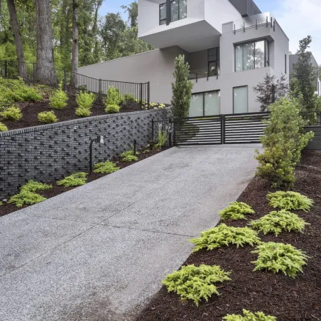 Modern white and gray house with a sloped driveway, black metal gate, and landscaped yard with green shrubs.