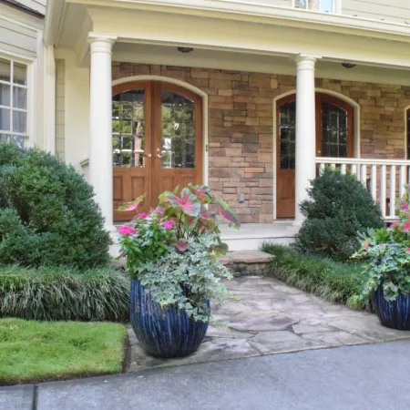 Front porch with stone walkway, large planters, green shrubs, and wooden double doors on a suburban home