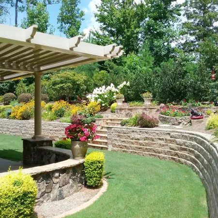Sunny backyard with stone retaining walls, colorful flowers, green lawn, and covered patio seating area.