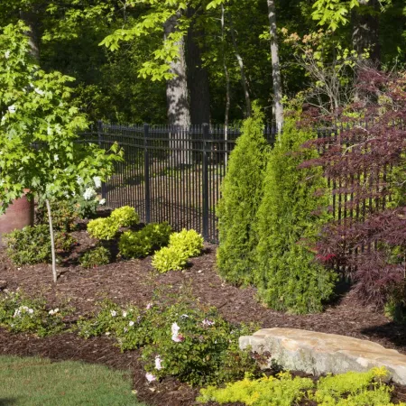 Well-maintained garden with shrubs, small trees, mulch beds, and a stone bench by a black iron fence under tall trees.