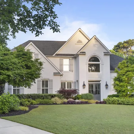 White two-story house with landscaped front yard, green lawn, and mature trees under a clear sky.
