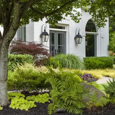 Lush garden with ferns, shrubs, and trees in front of a white house with lanterns and arched windows