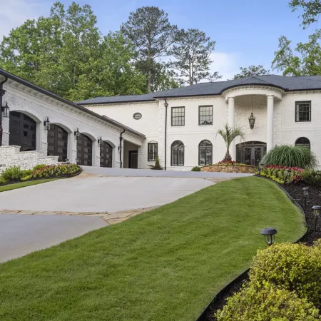 Large white brick mansion with black roof, arched windows, three-car garage, manicured lawn, and landscaped garden beds.