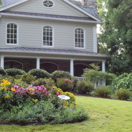 Two-story house with a manicured lawn and colorful flower garden in front surrounded by greenery.