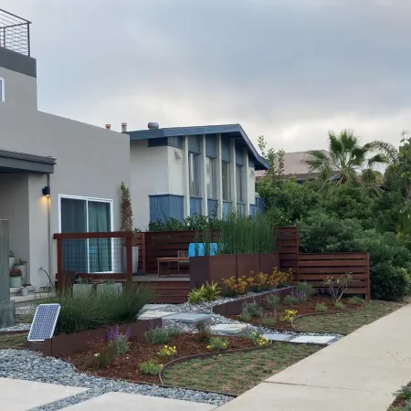Modern house with landscaped front yard featuring gravel, plants, wooden deck, and paved sidewalk on cloudy day