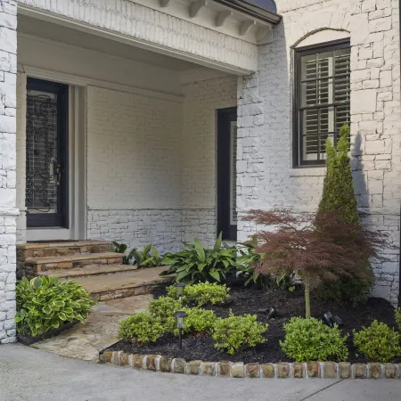 White brick house entrance with stone steps, black door, manicured garden, and small tree by window.