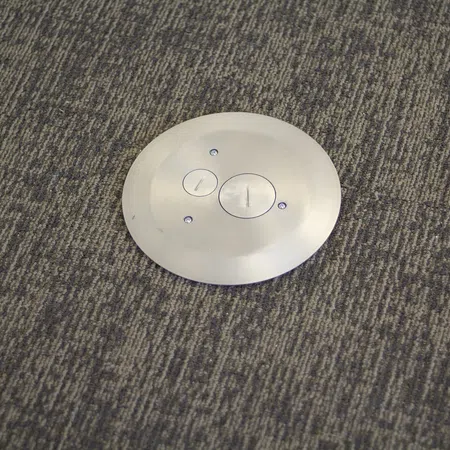 Round metal floor plate with two circular access covers installed on textured gray carpet flooring.