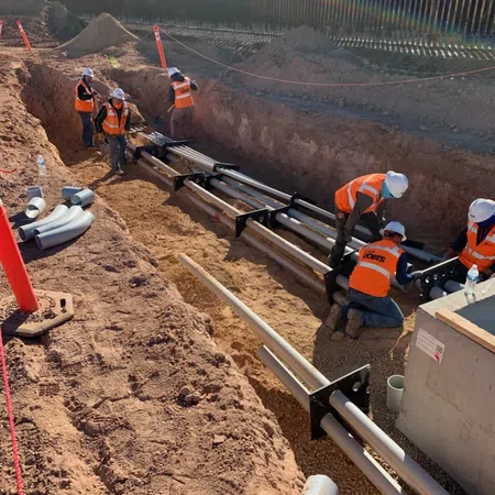 Construction workers in orange vests installing underground pipes in a trench near a metal fence under sunlight.