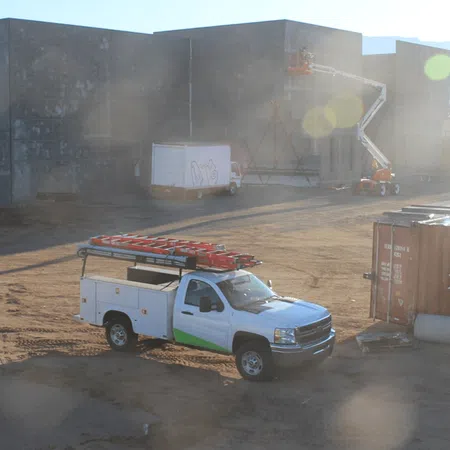 White utility truck with ladders parked on a dusty construction site near concrete buildings and a lift.