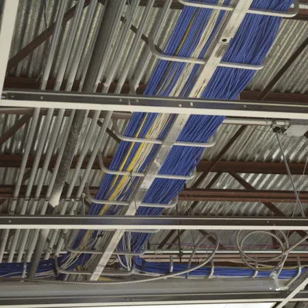 Blue and yellow cables organized in metal trays suspended from an industrial metal ceiling with pipes and beams.