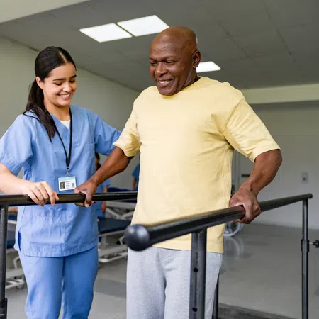 Nurse assisting elderly man with walking bars during physical therapy session in rehabilitation clinic.