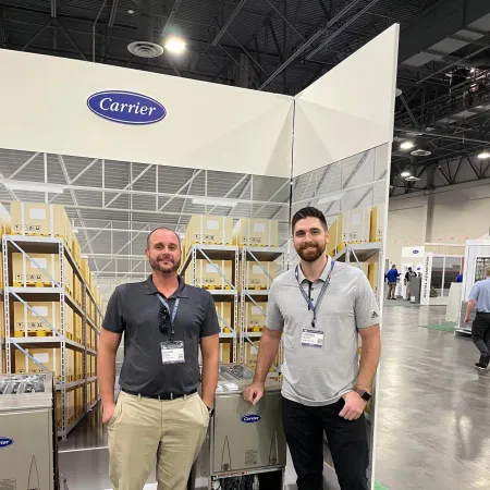 Two men standing beside Carrier HVAC units at a trade show booth with industrial shelving backdrop.