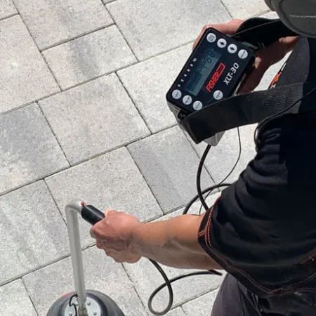 Person using a metal detector device on a paved stone surface with headphones on outdoors.