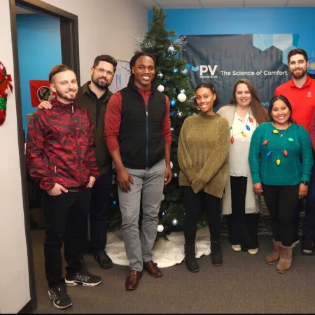 Group of eight diverse coworkers standing by a decorated Christmas tree in an office festive setting.