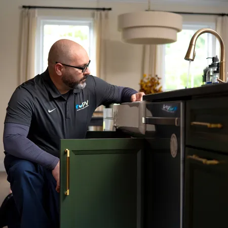 Technician inspecting plumbing under kitchen sink with green cabinets and gold handles in a modern home.