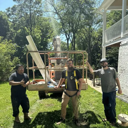 Three construction workers standing on grass next to equipment and a house on a sunny day