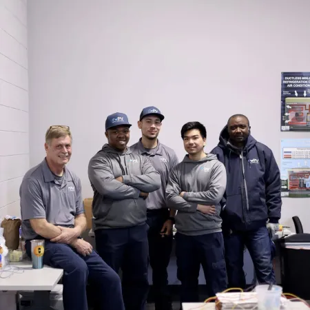 Five men in gray and navy workwear posing together in a workshop or office environment with equipment and posters.
