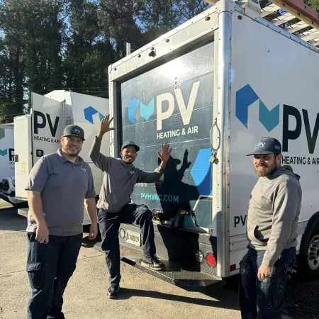 Three PV Heating & Air technicians in uniform posing happily by company service trucks outdoors.