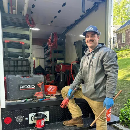 Smiling tradesman in workwear holding tools by a van loaded with toolboxes and equipment on a sunny day.