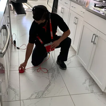 Technician with headphones using electronic equipment to inspect white kitchen floor tiles by cabinets and stove.