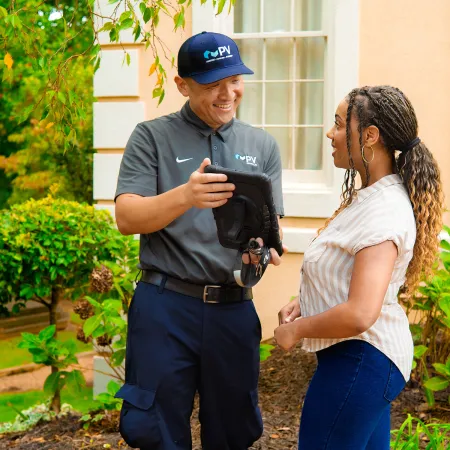 Technician in uniform showing a tablet to a woman outside a house with greenery and windows in the background