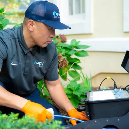 Technician in uniform and gloves using diagnostic equipment outdoors near a house wall and plants.