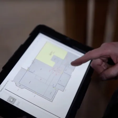 Person using a tablet to view and point at a digital floor plan layout in an indoor setting.