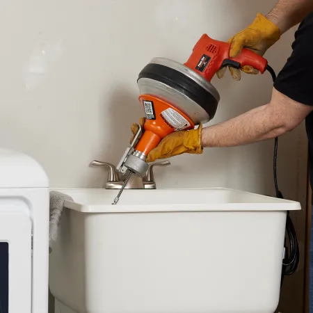 Worker using a power drain auger machine to clear a clogged sink drain in a utility room.