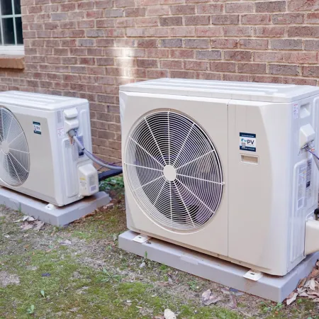 Two outdoor air conditioning units installed on concrete pads next to a brick house wall with wiring connected.
