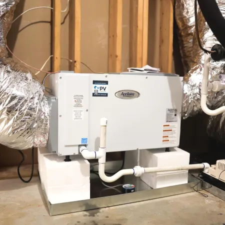 Aprilaire humidifier installed in a basement with PVC piping and exposed insulation ducts around wooden framing.