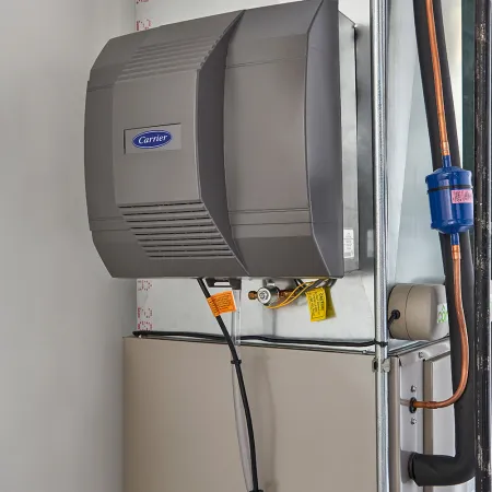 Carrier humidifier attached to home HVAC system with pipes and electrical wiring inside a utility room.