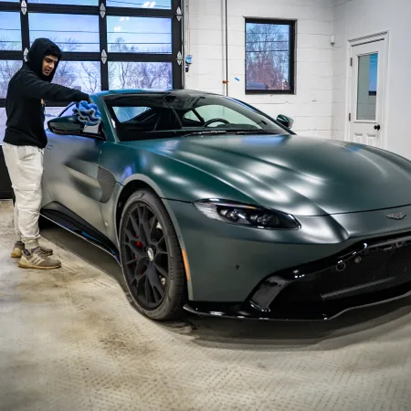 Person in hoodie polishing a dark green luxury sports car inside a garage with large windows and concrete floor
