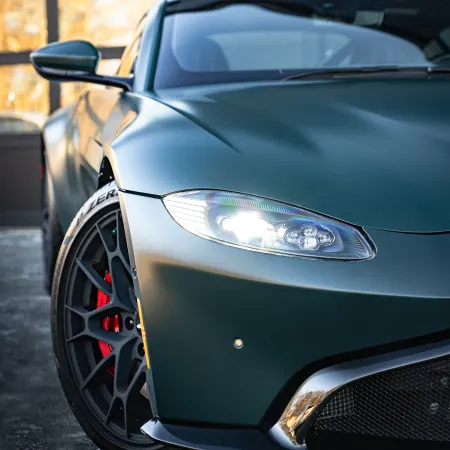Close-up of a sleek matte green sports car front with illuminated headlight and black alloy wheel.