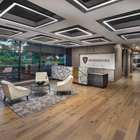 Modern orthodontics office reception with stylish chairs, marble desk, wood flooring, and geometric ceiling lights.