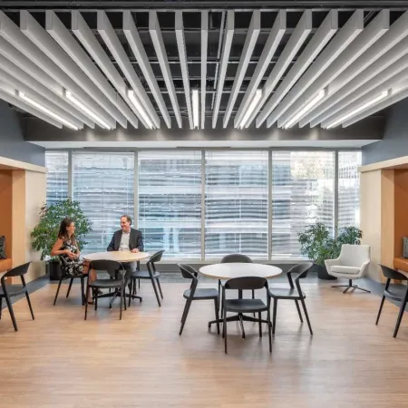 Modern office lounge area with wood flooring, built-in seating pods, round tables, and large windows lighting the space.