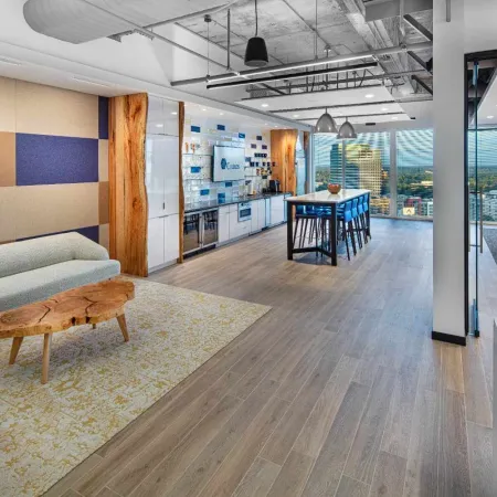 Modern office lounge with wooden coffee table, gray couches, kitchenette, high table, and glass-walled meeting room.