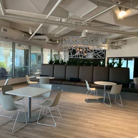 Modern office lounge with gray chairs, round tables, wood flooring, and large windows allowing natural light.